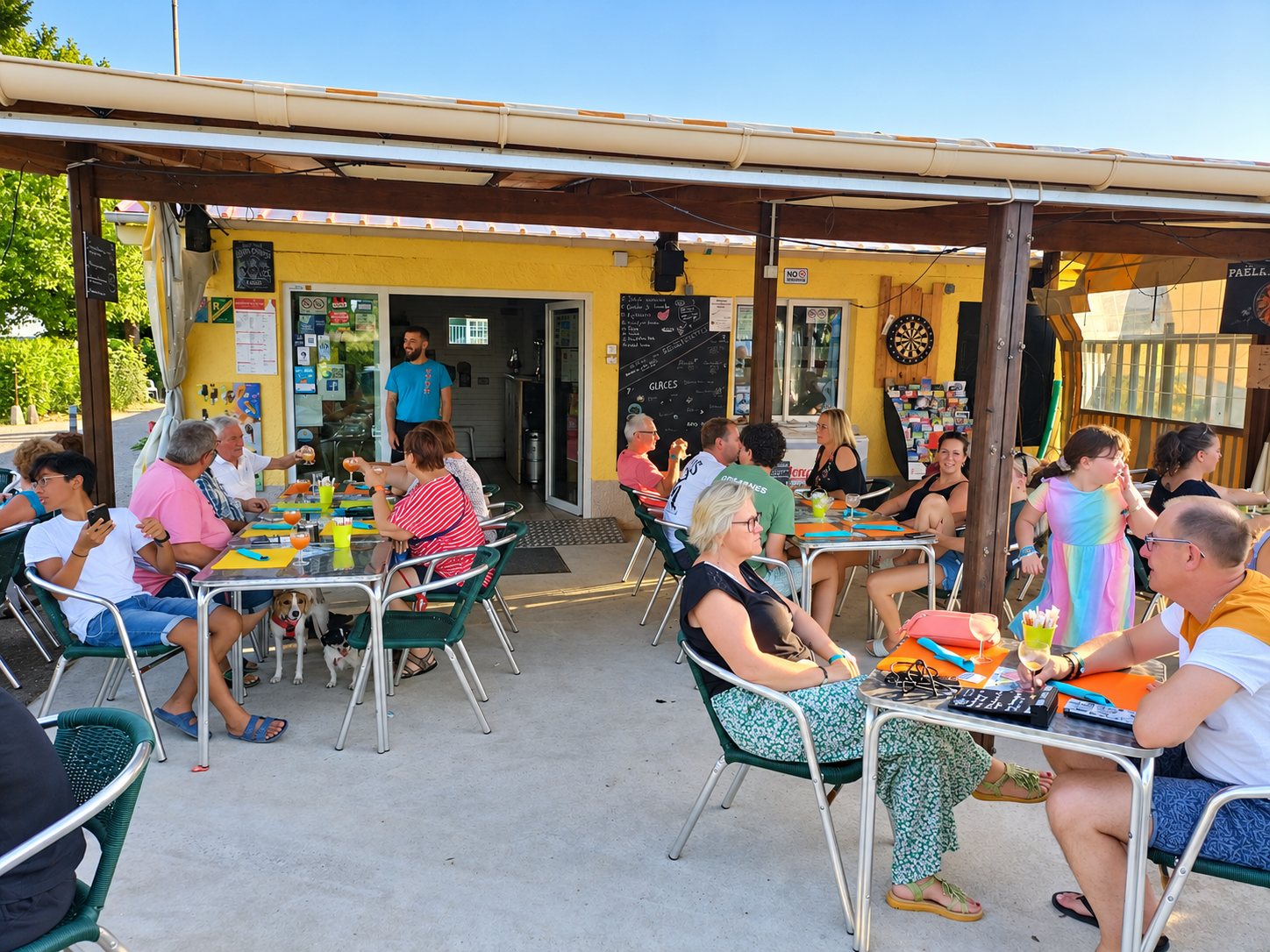 Des clients installés à des tables sur une terrasse couverte devant un bar jaune, avec un serveur debout à l'entrée et des chiens sous les tables.
