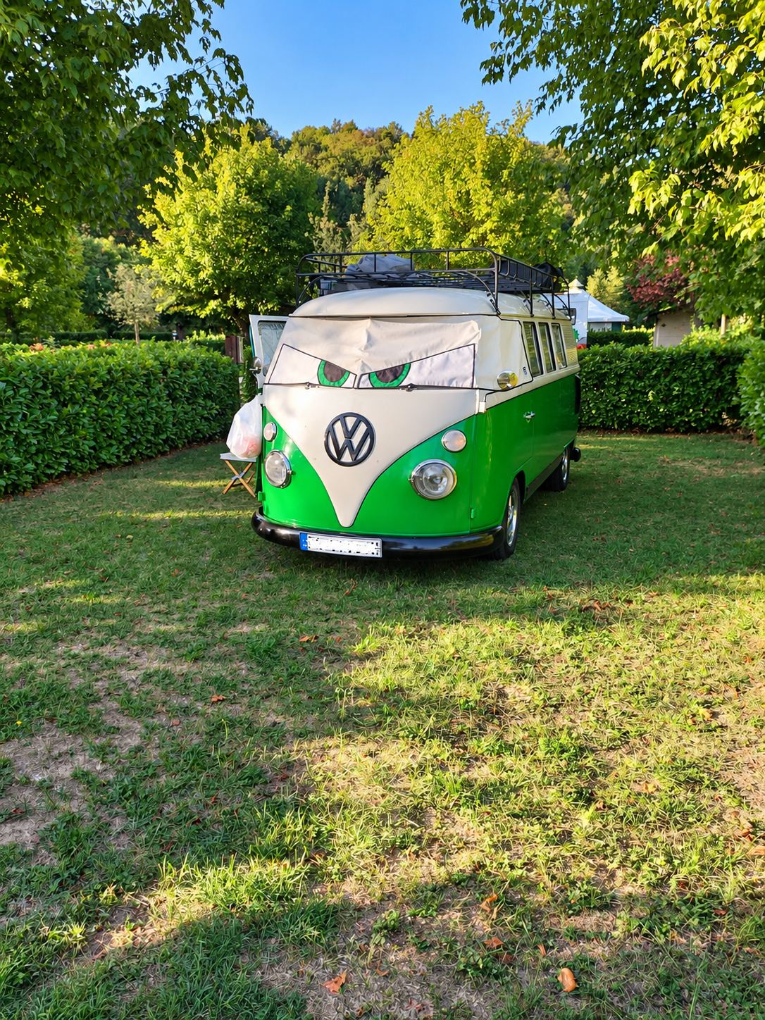 Un combi Volkswagen vintage vert et blanc, avec une galerie de toit et un cache-pare-brise décoré de grands yeux, est stationné sur une pelouse entourée de haies et d'arbres.