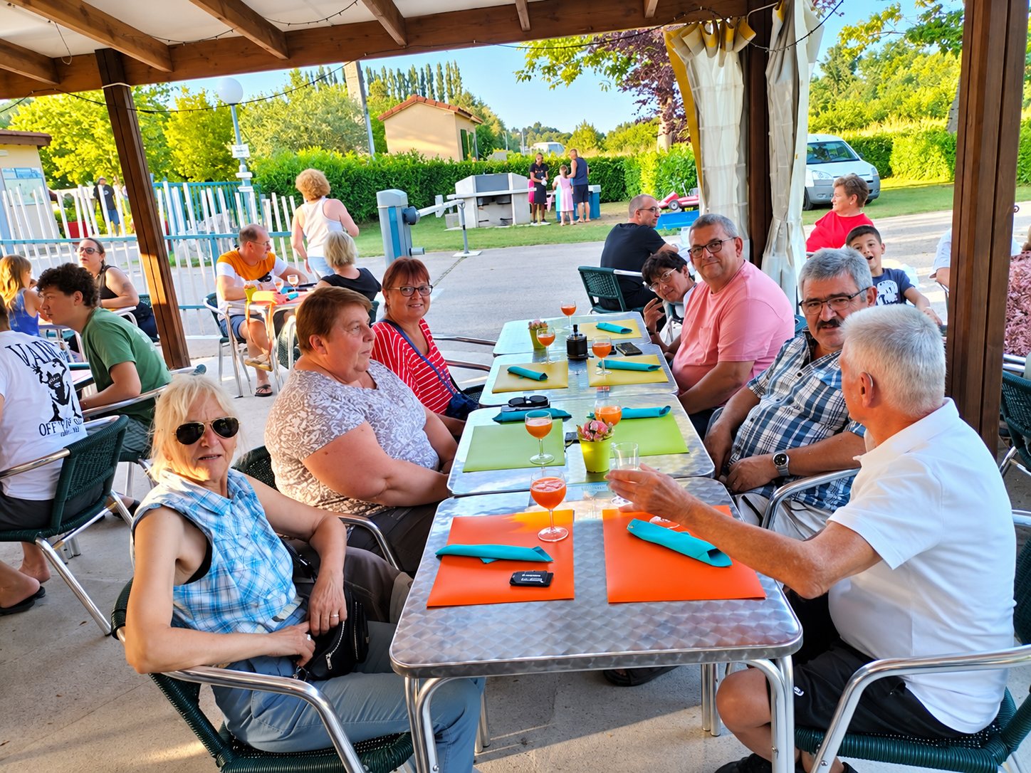 Des convives souriants sont assis autour de tables en métal avec des sets de table colorés et des verres d'apéritif sous une terrasse couverte, par une soirée ensoleillée.