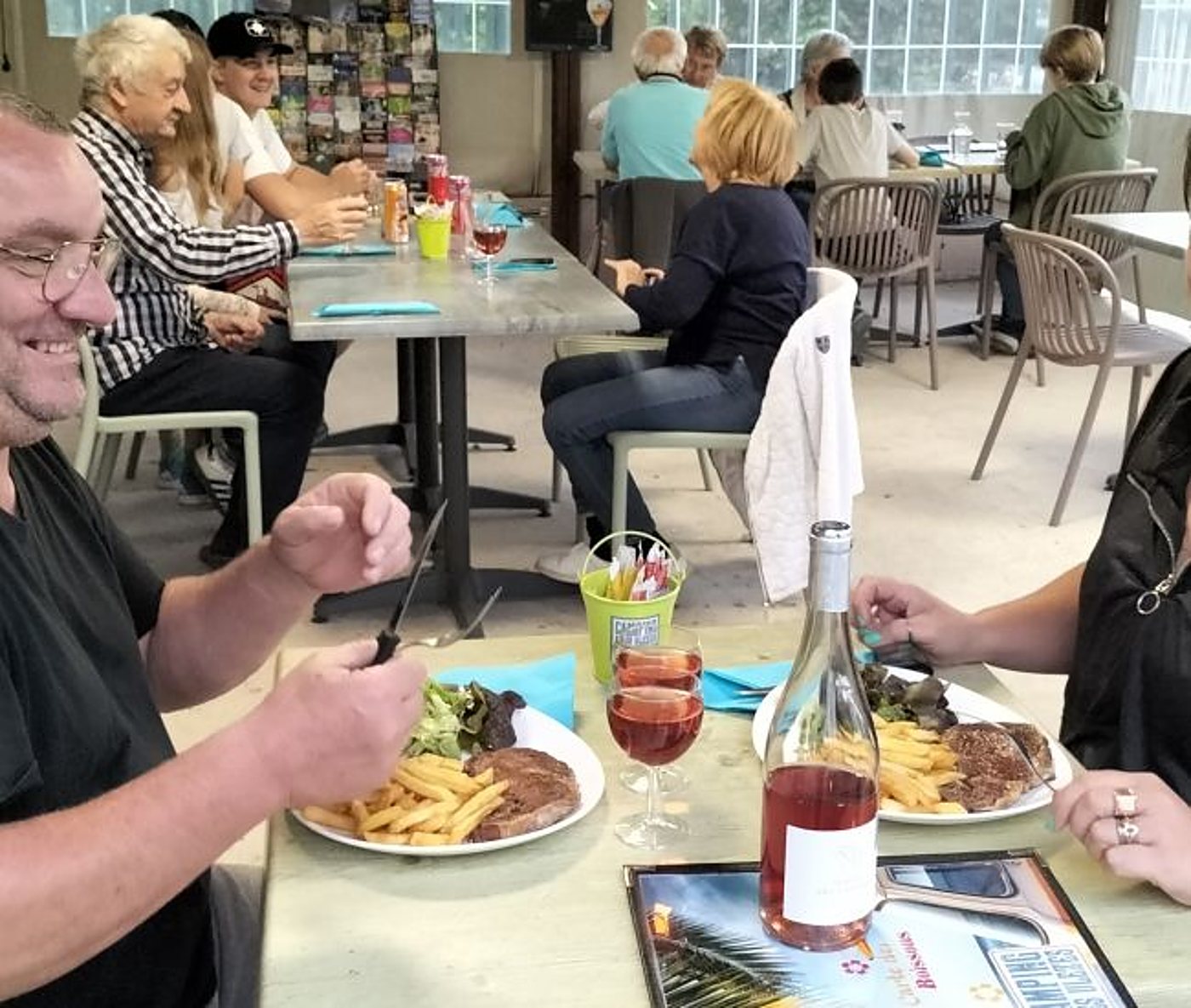 Des clients attablés dans une salle de restaurant partagent un repas de steak frites accompagné de vin rosé, avec d'autres tables occupées en arrière-plan.
