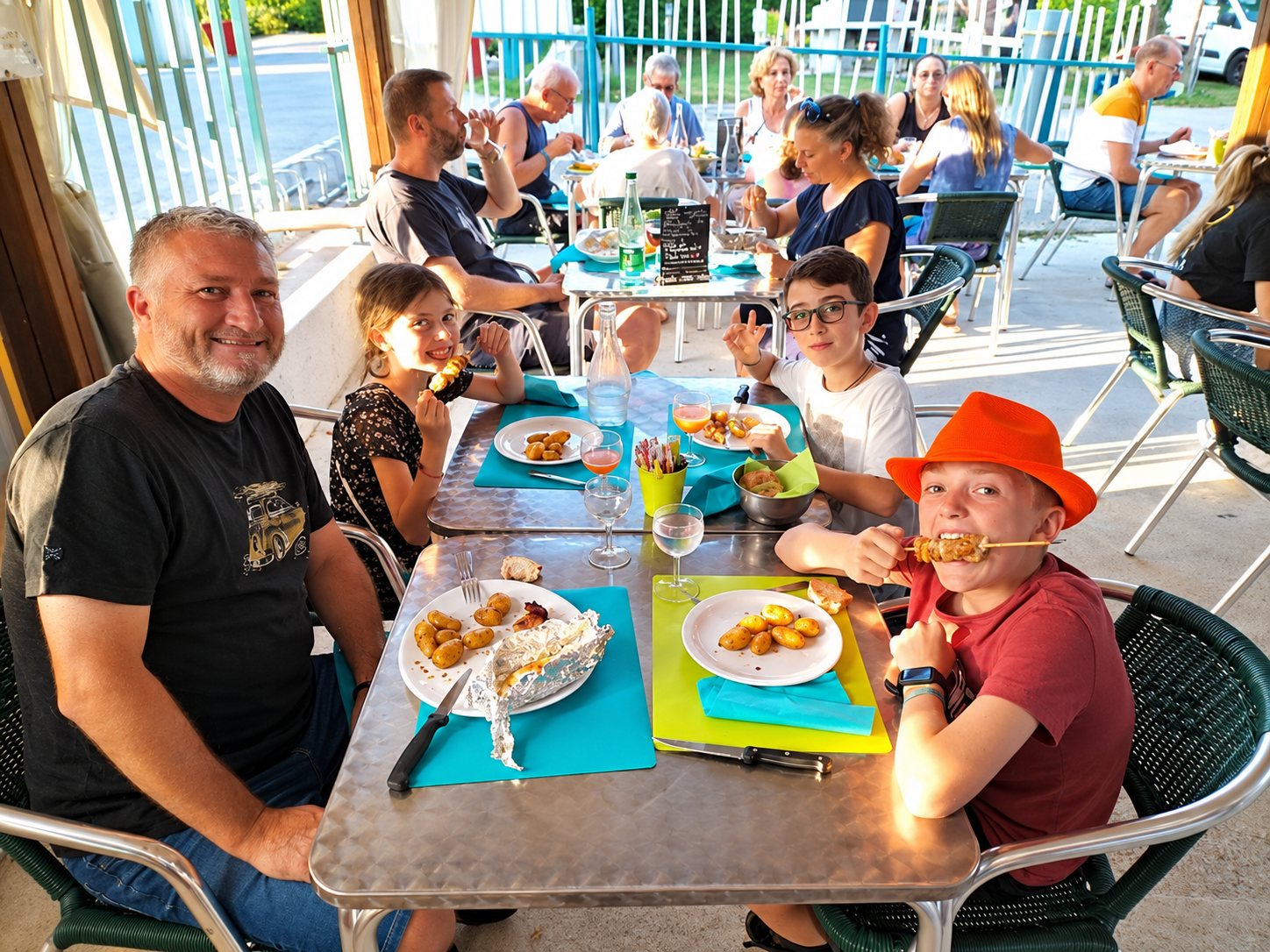 Un homme et trois enfants sourient à une table de terrasse en inox où sont servis des brochettes et des pommes de terre, avec d'autres convives en arrière-plan.