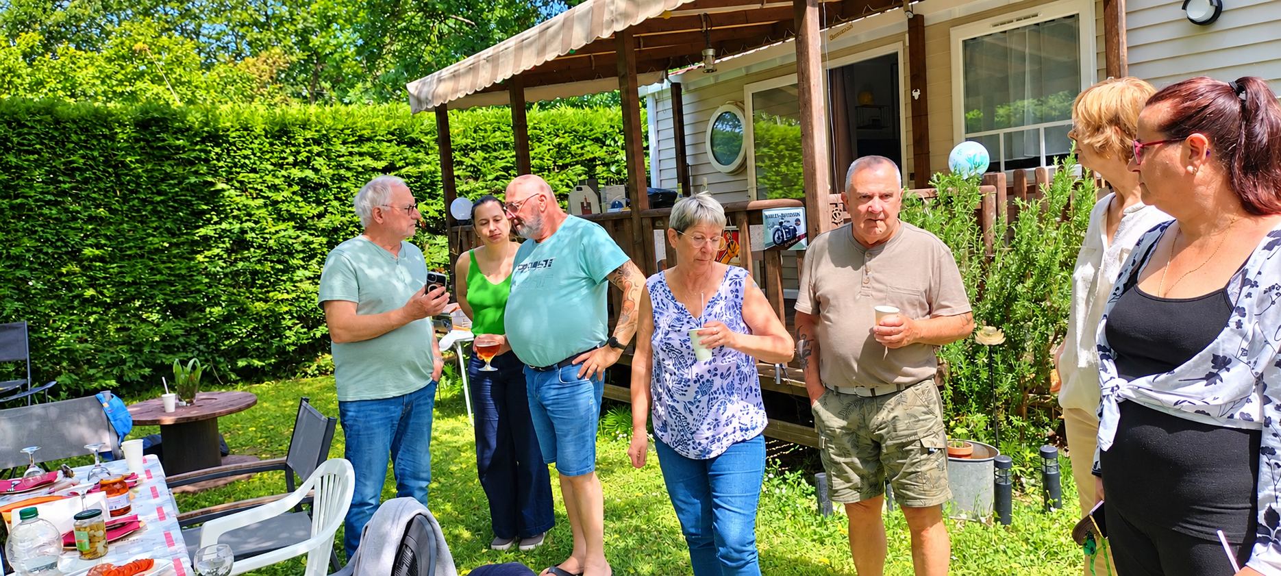 Un groupe de personnes discute un verre à la main dans un jardin devant un mobil-home, près d'une table dressée.