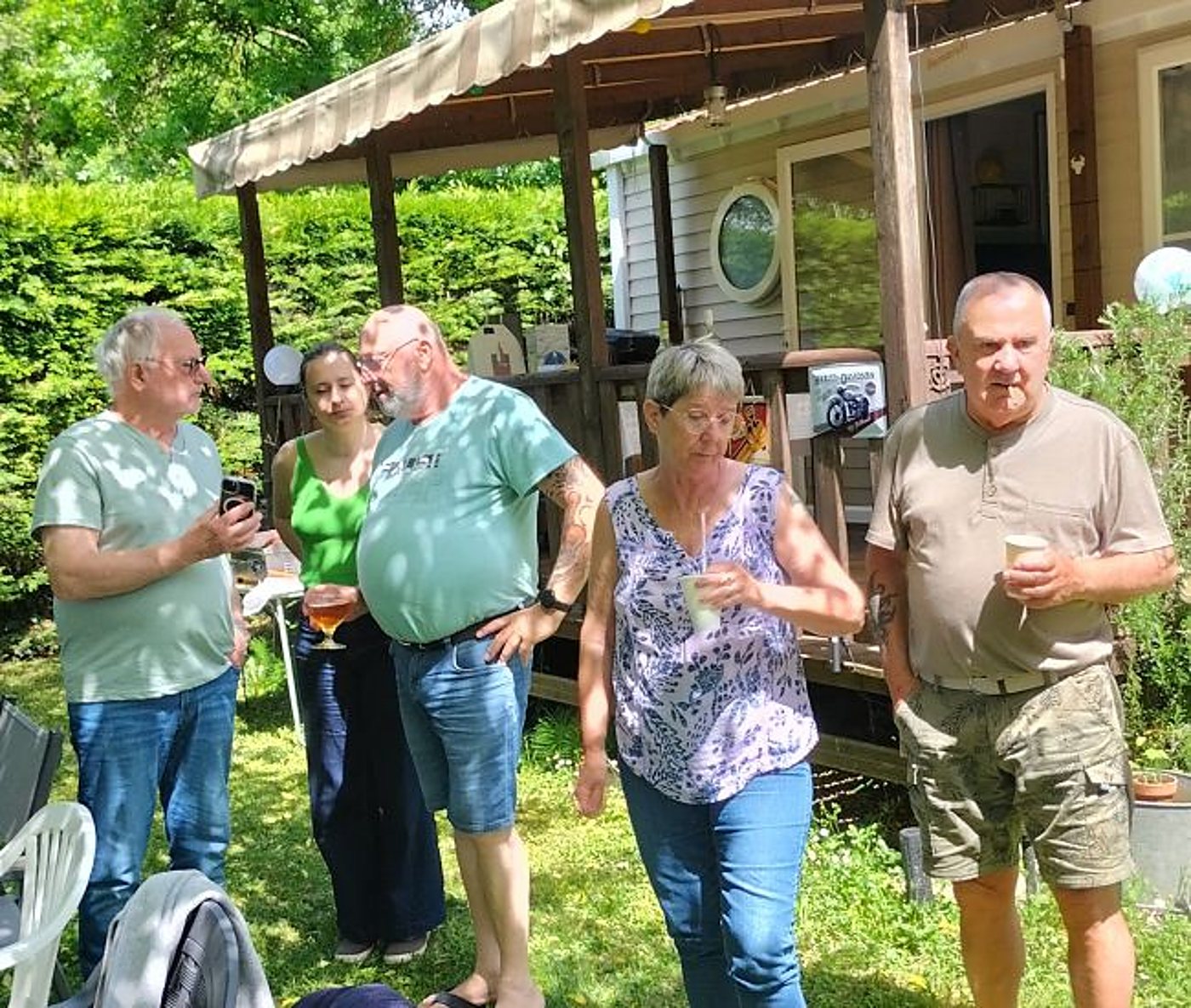Cinq personnes se tiennent debout sur l'herbe, verres à la main, devant un mobil-home avec une terrasse couverte en bois, par une journée ensoleillée.