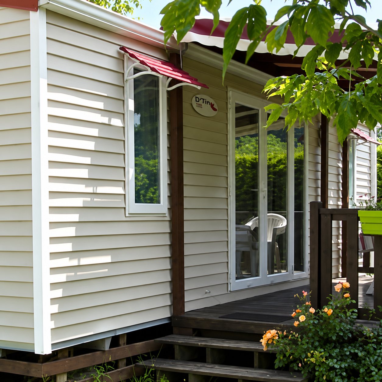 Un mobil-home au bardage beige clair avec une petite terrasse en bois, une baie vitrée et des fleurs au premier plan, sous le feuillage d'un arbre.