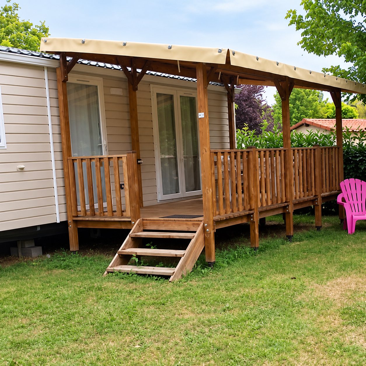 Un mobil-home beige équipé d’une terrasse en bois avec garde-corps et toit en toile, installé sur une pelouse, avec une chaise rose au bord.