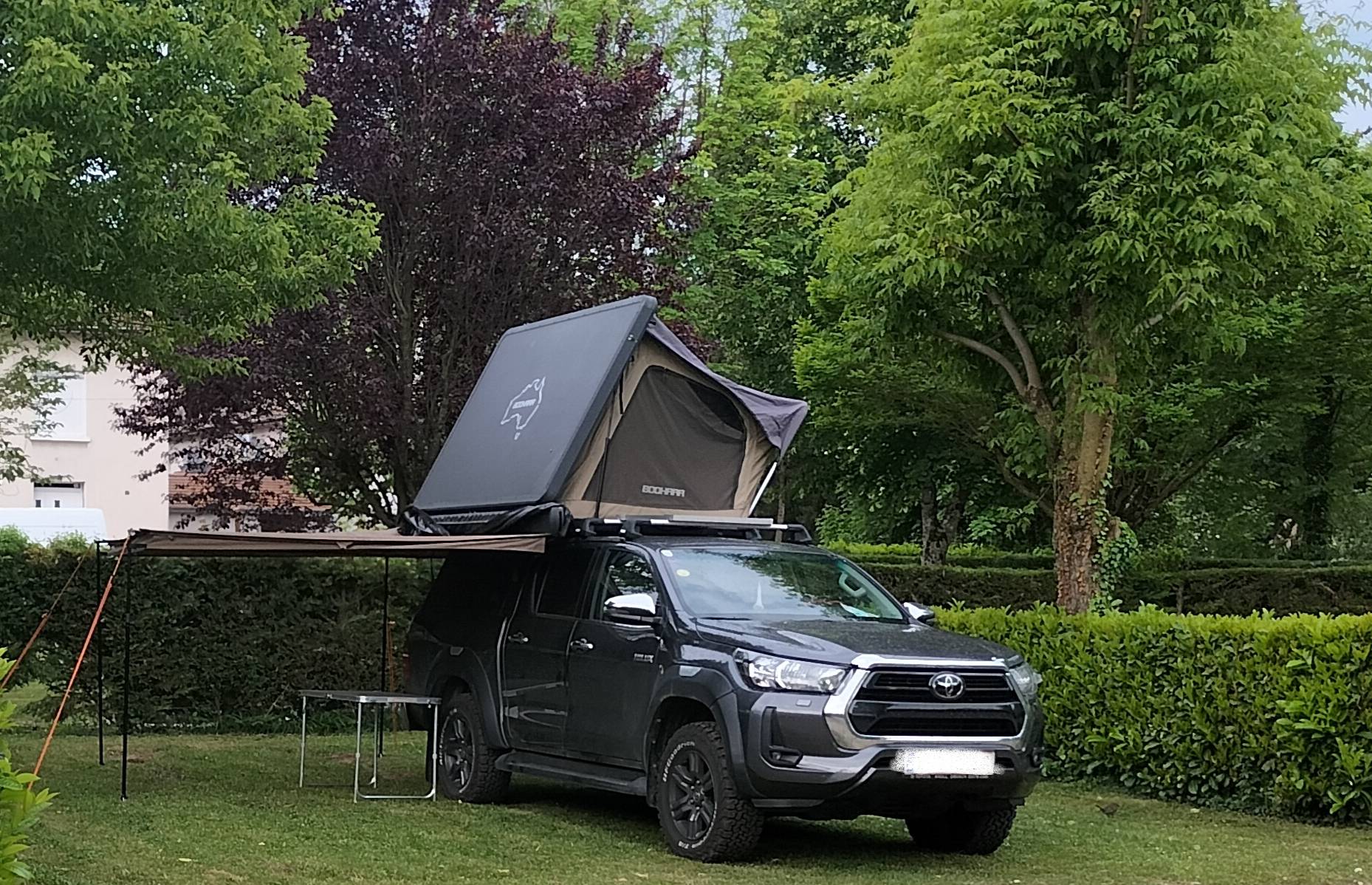 Un pick-up Toyota gris foncé garé sur une pelouse porte une tente de toit ouverte et un auvent déployé, entouré d'arbres et de haies.