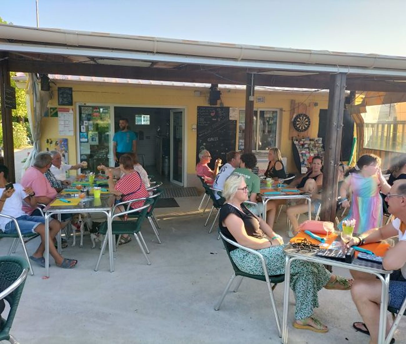 Des clients assis à des tables métalliques sur la terrasse couverte d'un bar aux murs jaunes, avec une cible de fléchettes et un serveur à l'entrée.